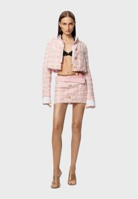 Giacca cropped in tweed rosa con bottoni dorati abbinata a una gonna coordinata. Polsini bianchi e top nero sotto. Tacchi trasparenti completano il look.