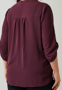 Femme portant un haut à manches longues de couleur violet foncé avec des détails de tissu froncé sur le haut du dos, se tenant devant un fond uni.