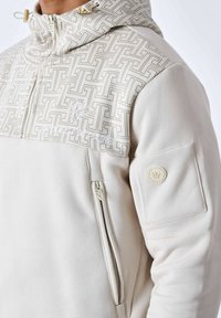Hoodie beige clair avec fermeture éclair, présentant un empiècement texturé et des poches sur les côtés. Comprend un détail de logo sur le bras.