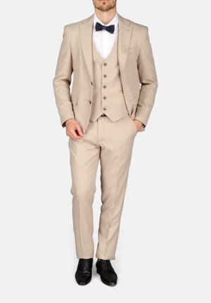 Man in beige driedelig pak met wit overhemd, zwarte vlinderdas en zwarte nette schoenen, staand met één hand in de zak.