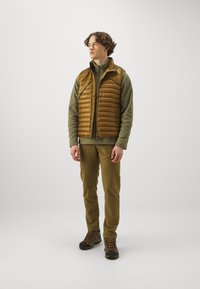 Brown puffy vest over olive fleece, paired with olive pants and brown hiking boots, featuring textured fabric and zipper details.