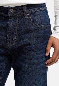 Dunkelblaue Jeans aus Denim mit schmaler Passform, klassischem Fünf-Taschen-Design und kontrastierenden gelben Nähten. Der Stoff hat eine glatte Textur.