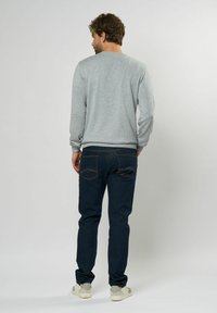 Grijze sweatshirt met ronde hals, donkerblauwe jeans met oranje stiksels, en witte sneakers, gezien van achteren tegen een lichte achtergrond.