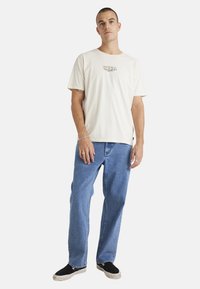 Light beige cotton T-shirt with a graphic logo on the chest, paired with blue straight-leg jeans and black slip-on shoes.