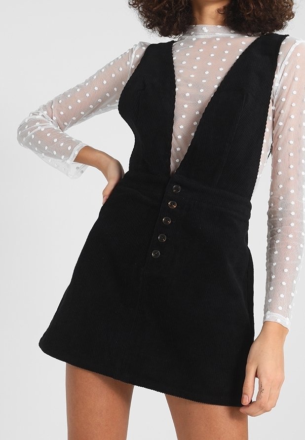 Nainen, joka käyttää mustaa corduroy-materiaalia olevaa hihattomaa mekkoa, jossa on etunapit, valkoisen läpikuultavan pilkkupaidan päällä, käsi lantiolla.