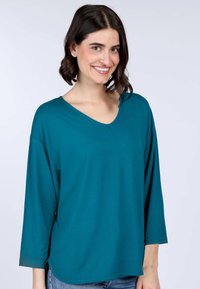 Lockeres V-Ausschnitt-Oberteil in Petrol mit Dreiviertelärmeln, weichem Stoff und geradem Saum, kombiniert mit hellblauen Jeans.