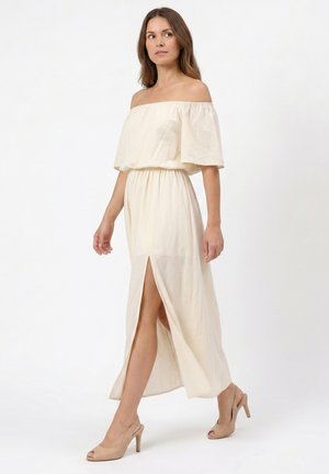 Vrouw wandelend in een crèmekleurige off-shoulder jurk met een voorste split, gecombineerd met nude open-toe pumps met hoge hak, tegen een witte achtergrond.
