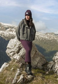 Grau gestrickte Jacke mit zwei vorderen Taschen, lila knielanger Rock und schwarzen Knöchelstiefeln. In einer bergigen Landschaft im Freien.