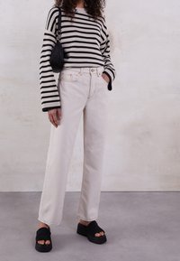 Pruhovaný černobílý pletený svetr, krémové džíny s vysokým pasem a černé platformové sandály. Obsahuje široké rukávy a třepený lem džínů.