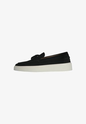 Black suede slip-on shoes with a rounded toe, tassel detail, and a white thick sole. Features stitching along the sole.