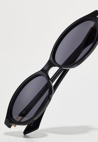 Black sunglasses with oval lenses, dark gray tint, and glossy plastic frames. Features slender temples and subtle branding on left arm.