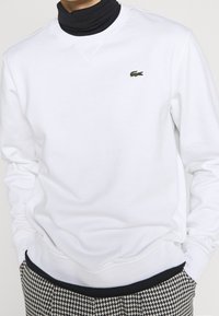 Sudadera blanca con cuello redondo y mangas largas, con un pequeño logo bordado de cocodrilo verde en el pecho. Corte casual.