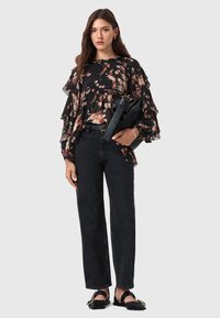 Blusa nera con stampe floreali e maniche a volant, abbinata a jeans neri a vita alta e sandali neri flat. La modella tiene una grande borsa nera.