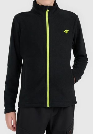 Enfant portant une veste polaire noire avec une fermeture éclair vert néon et un logo 4F, debout avec une main dans la poche devant un fond uni.