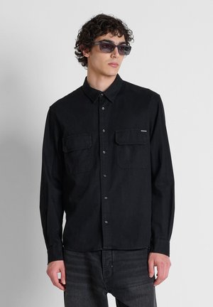 Joven con camisa negra abotonada con bolsillos en el pecho y gafas de sol oscuras, de pie frente a un fondo liso de color gris claro.