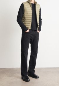 Gilet imbottito oliva sopra un maglione nero, abbinato a jeans neri a gamba dritta e stivali neri, in piedi su uno sfondo neutro.