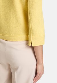 Žlutý svetr z filcu s texturovaným povrchem, s tříčtvrtečními rukávy a rovnými lemy se postranními rozparky, doplněný o světlé kalhoty.