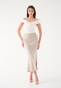 Blusa bianca a spalle scoperte con tessuto arricciato e dettaglio a rosa, abbinata a una gonna in satin beige. La modella indossa tacchi nudi con laccetti.