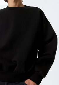 Juodas džemperis su apvaliu iškirpimu, laisvo silueto ir ribotomis rankogaliais. Minkšta tekstūra su lygiu paviršiumi, su juostele apatinėje dalyje.