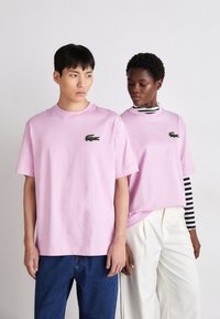 Due modelli indossano T-shirt Lacoste di un viola chiaro; uno le abbina a jeans blu, l'altro a pantaloni bianchi e a una maglietta a righe a maniche lunghe.