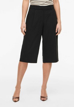 Nő fekete, térdig érő, bőszárú rövidnadrágot, csíkos berakott felsőt és barna, nyitott orrú magassarkút visel, fehér háttér előtt áll.