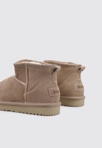 Botines tobilleros de ante color canela con un forro mullido, suela plana de goma y detalles con costuras. Incluyen una etiqueta en la parte trasera y una lengüeta para facilitar su colocación.