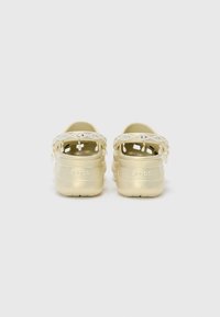 Gold Crocs with a beaded strap featuring dangling accents, smooth synthetic material, open back, and textured soles for grip.