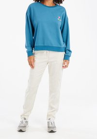 Felpa di cotone blu con scollo rotondo e texture morbida, caratterizzata da una piccola grafica multicolore sul petto. Abbinata a pantaloni beige chiaro.