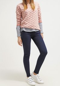 Felpa rosa a trama con punti in rilievo, sovrapposta a una camicia blu a fantasia, abbinata a jeans skinny scuri e sneakers bianche.