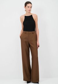 Musta pleated halterneck-top metalliketjuyksityiskohtien kanssa; yhdistetty laajoihin ruskeisiin housuihin; malli poseeraa kädet taskuissa; sileät kankaat.