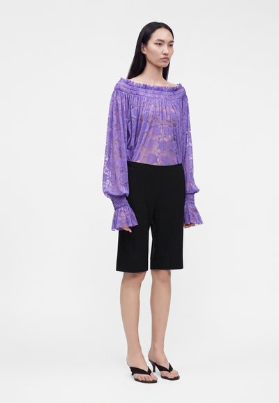 Femme portant un chemisier violet transparent à épaules dénudées avec manches longues, un short noir au genou et des sandales à talons noires, debout sur un fond blanc.
