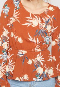 Rostorange Bluse mit floralem Druck in Weiß, Blau und Grün. Sie hat lange Ärmel und eine taillierte Form mit Peplum-Details.