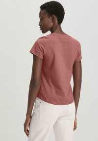 Lyhythihainen, istuva T-paita pölyruusun värissä, valmistettu puuvillasta. Siinä on pyöreä kaula-aukko ja kaareva helma, yhdistettynä vaaleisiin housuihin.