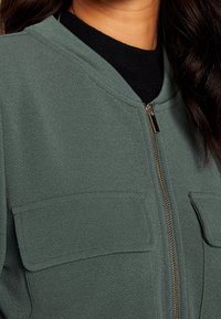 Giacca verde con zip in tessuto testurizzato, dotata di due tasche sul petto e colletto a costine. La cerniera dorata aggiunge un dettaglio a contrasto.