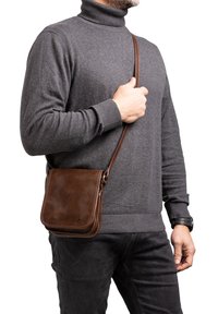 Borsa a spalla in pelle marrone con una forma quadrata, caratterizzata da una texture liscia e una lunga tracolla, indossata da un uomo con un maglione a collo alto grigio.