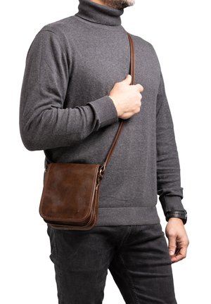 Borsa a spalla in pelle marrone con una forma quadrata, caratterizzata da una texture liscia e una lunga tracolla, indossata da un uomo con un maglione a collo alto grigio.