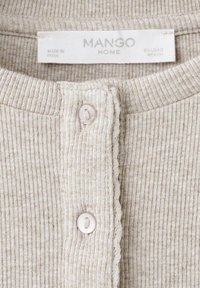Gros plan sur une chemise en tissu côtelé beige avec deux boutons blancs et une étiquette indiquant Mango Home, fabriquée en Inde, taille EU USA S MEX CH.