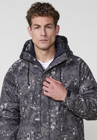 Chaqueta acolchada en un patrón gris oscuro con detalles en blanco. Cuenta con una capucha, cierre frontal y puños elásticos. El material parece texturizado.