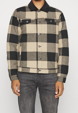 Mand iført beige og sort ternet skjortejakke med sort krave og mørkegrå jeans mod en ensfarvet baggrund.