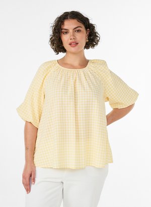 Donna con capelli corti e ricci che indossa una camicetta gialla a quadri e pantaloni bianchi, posando con una mano sui fianchi davanti a uno sfondo bianco.