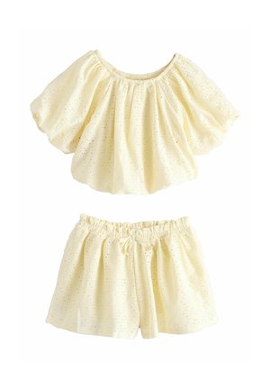 To-delt sæt i cremefarvet hulmønsterstof med puffede kortærmede top og elastik i talje på shorts med lille sløjfe foran.
