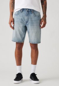 Șorturi din denim de culoare albastră deschisă, cu o croială lejeră, cinci buzunare și margine desfăcută. Asortate cu adidași negri și șosete albe până la gleznă.