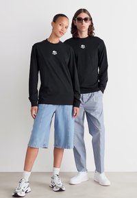Zwei Modelle tragen schwarze Langarmshirts von Lacoste; eines in hellblauen knielangen Shorts und Sneakers, das andere in hellblauen Hosen und weißen Sneakers.