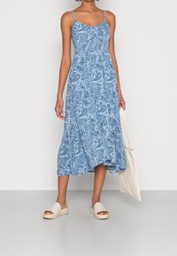 Modré šaty s paisley vzorem, tenkými ramínky, střední délkou a volnou siluetou. Doplněné béžovými sandály na plochém podpatku a taškou.