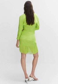 Grünes Satin-Kleid mit langen Ärmeln und Fransenverzierung am Saum. Getragen mit weißen Riemchensandalen, die eine figurbetonte Silhouette betonen.
