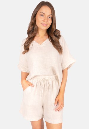 Young woman with long brown hair wearing a loose beige short-sleeve top and matching drawstring shorts, standing with one hand in pocket.