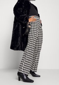 Svart pälsjacka, figursydd svart topp, vita byxor med herringbone-mönster och vida ben, samt svarta högklackade skor. Svart rund handväska.