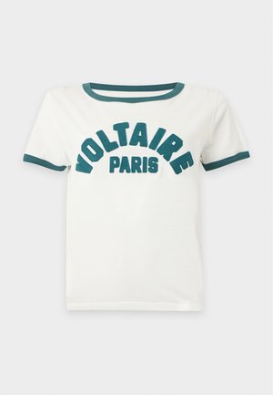 Femme portant un t-shirt blanc avec le texte "Voltaire Paris", une jupe en cuir marron, des bottes noires mi-mollet, tenant un sac à main noir clouté.