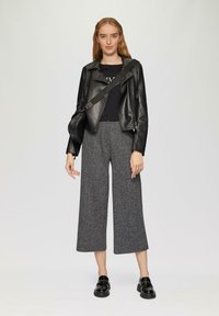 Schwarze Lederjacke, enganliegendes schwarzes Hemd, weite graue gemusterte Hose und schwarze Slipper. Eine einfache Tasche hängt über der Schulter.