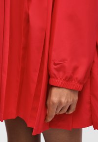 Χέρι ακουμπισμένο σε κόκκινη πλισέ φούστα, φορά κόκκινο μπουφάν με μακριά μανίκια, ελαστικό τελείωμα και μικρό λογότυπο Nike.